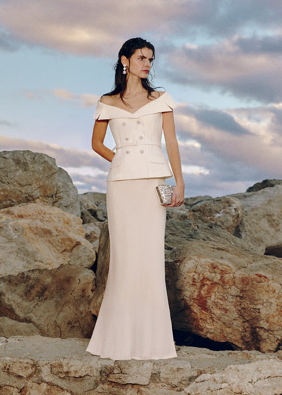 Woman in a white off-shoulder dress standing on rocky terrain with a cloudy sky.