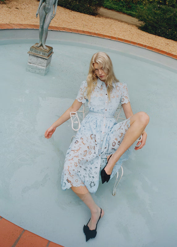 Woman in a blue lace dress sitting by a pool with a statue in the background.