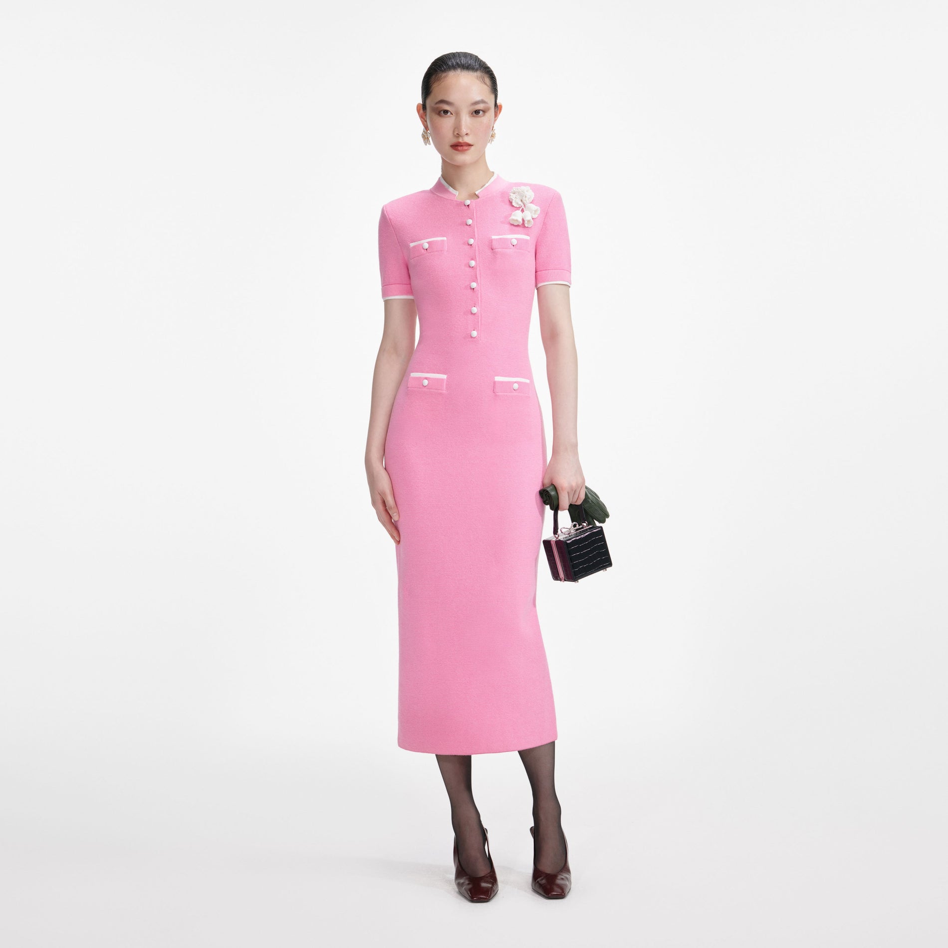 Woman in a pink midi dress with white trim, white buttons, and a white floral brooch, holding a black handbag
