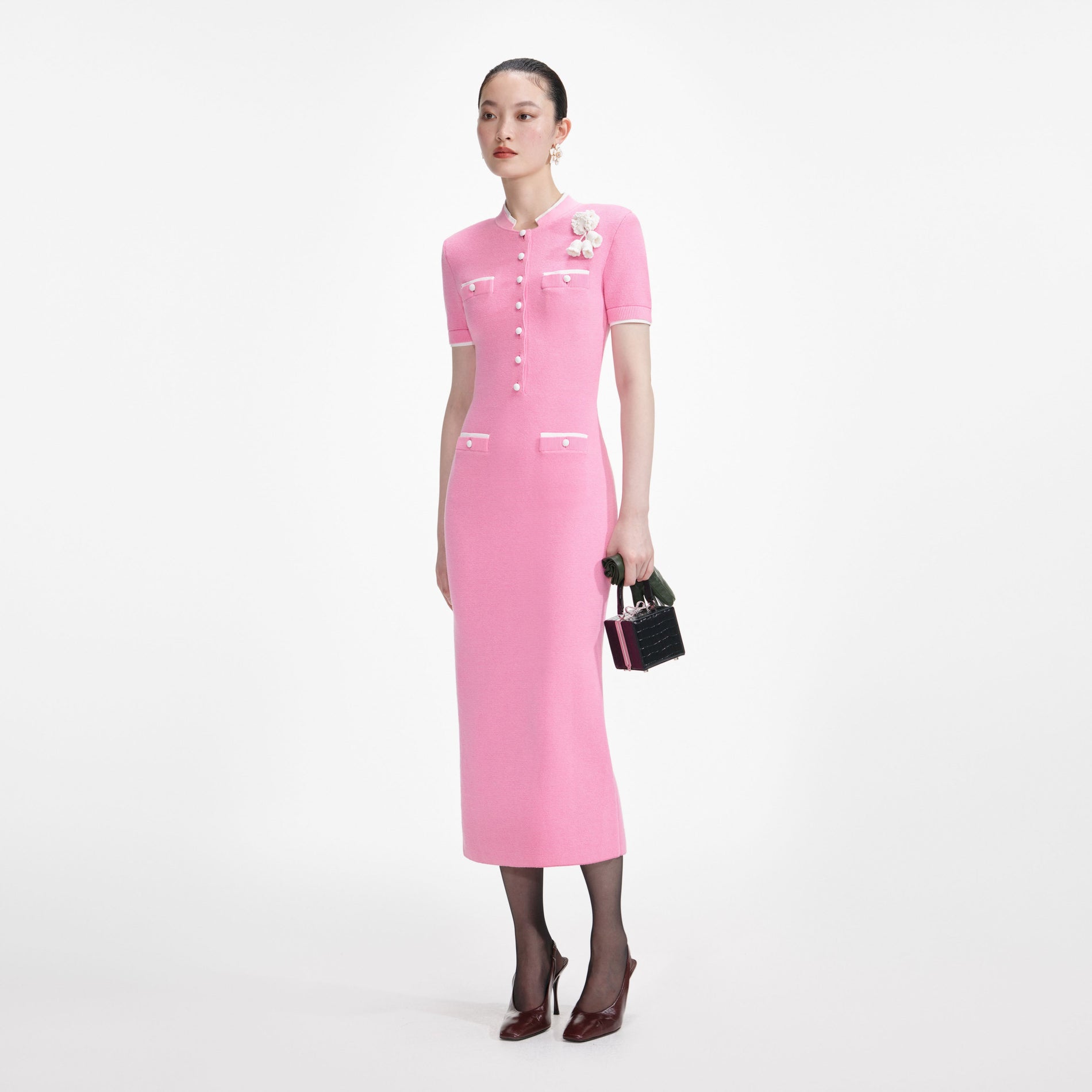 Woman in a pink midi dress with white trim, white buttons, and a white floral brooch, holding a black handbag