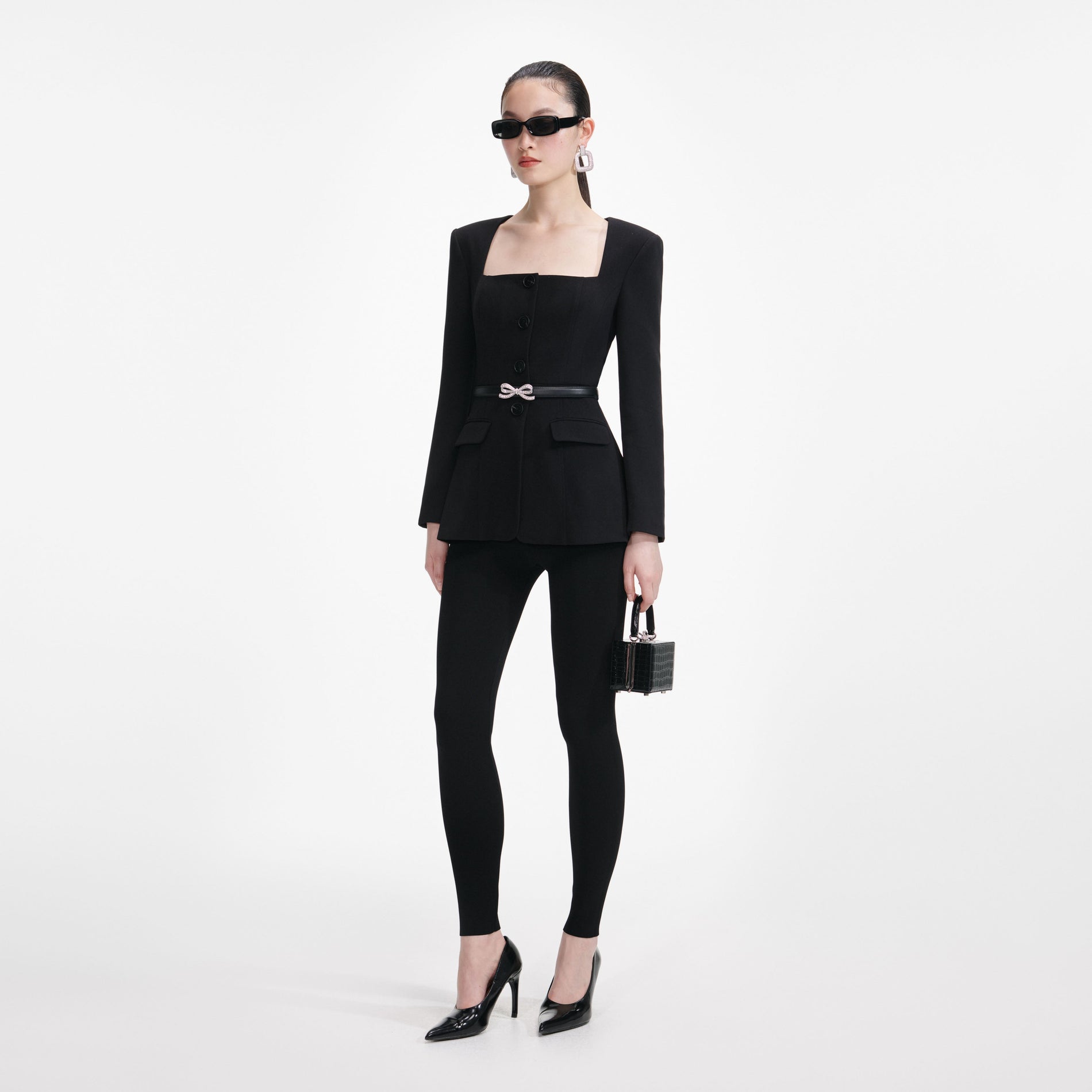 Woman in black square-neck blazer, leggings, sunglasses, and heels, holding a small black handbag