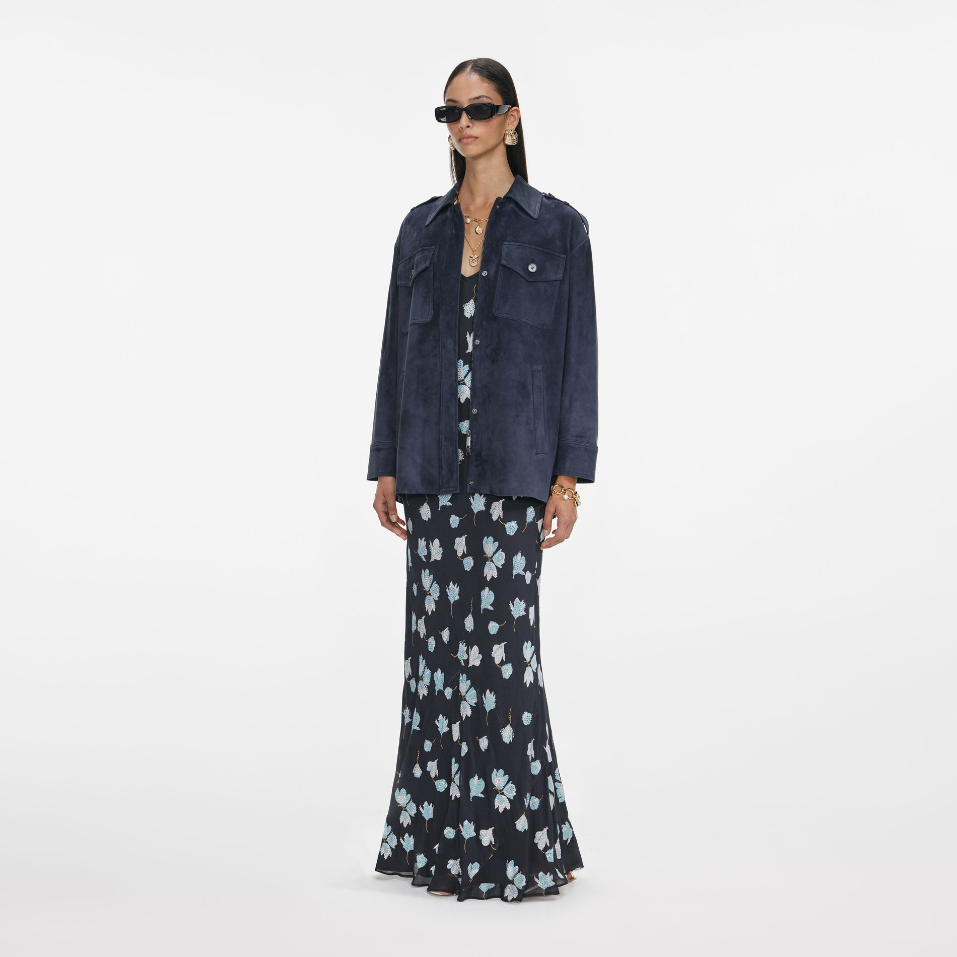 Woman in a navy blue suede jacket over a long floral dress, sunglasses, and gold jewelry