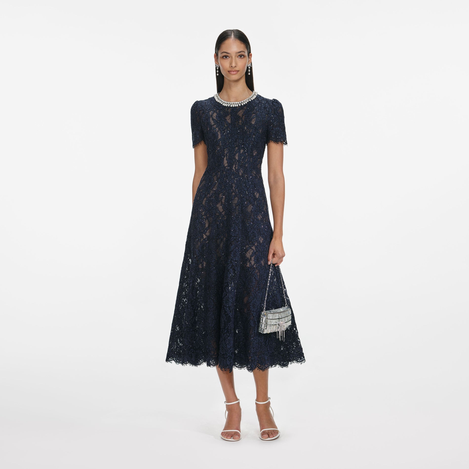 Woman in a navy blue lace midi dress with short sleeves, pearl embellished neckline, and white heels