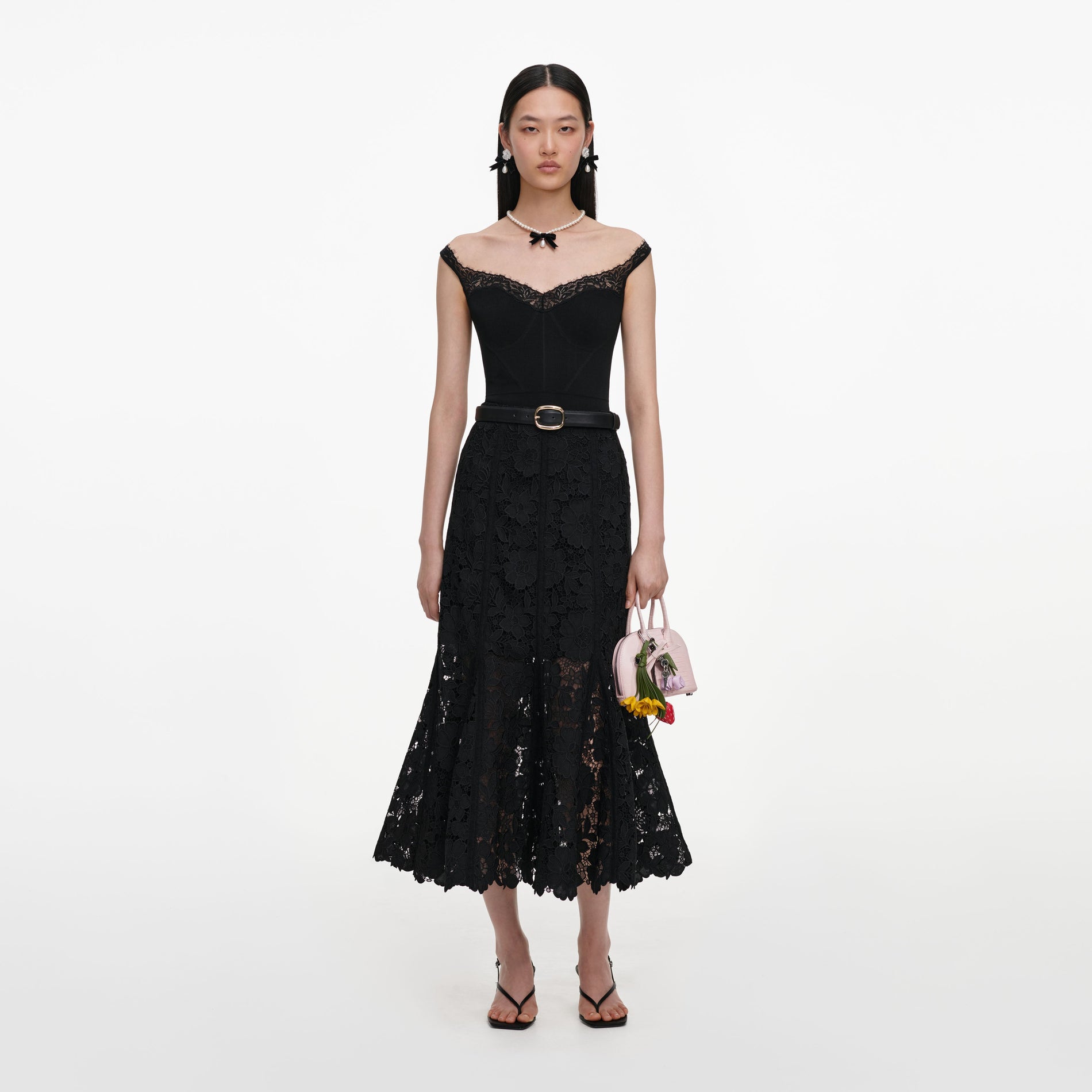 Woman in black off-shoulder lace dress with belt, pearl necklace, and earrings, holding a pink handbag