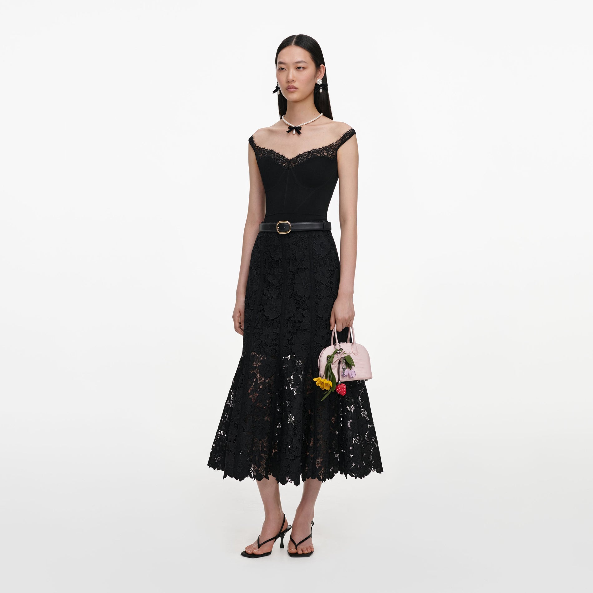 Model in black lace dress, pearl necklace, and black heels, holding a pink handbag with flower charms