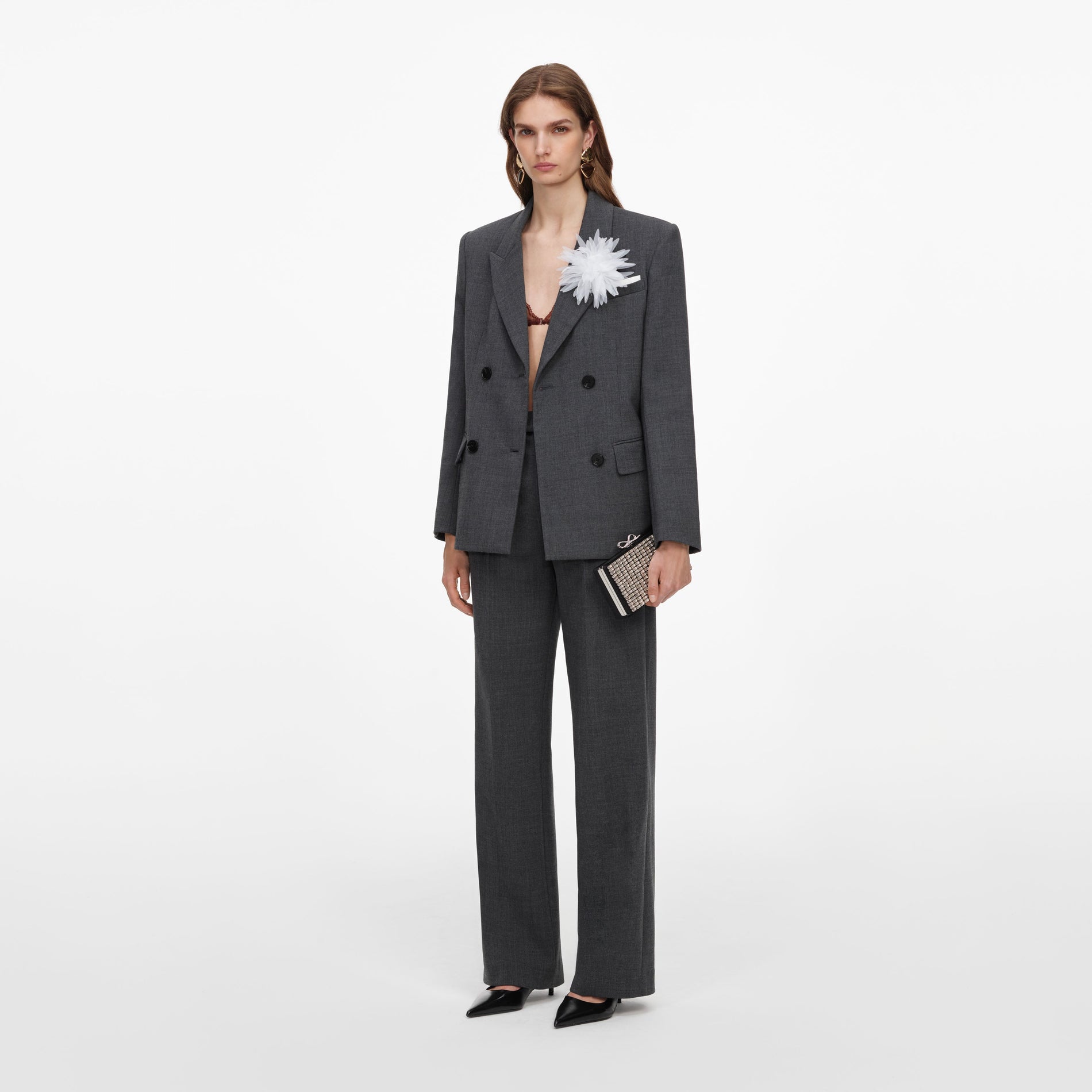 Woman in a dark grey double-breasted suit, with a white flower pinned to the lapel, holding a clutch bag