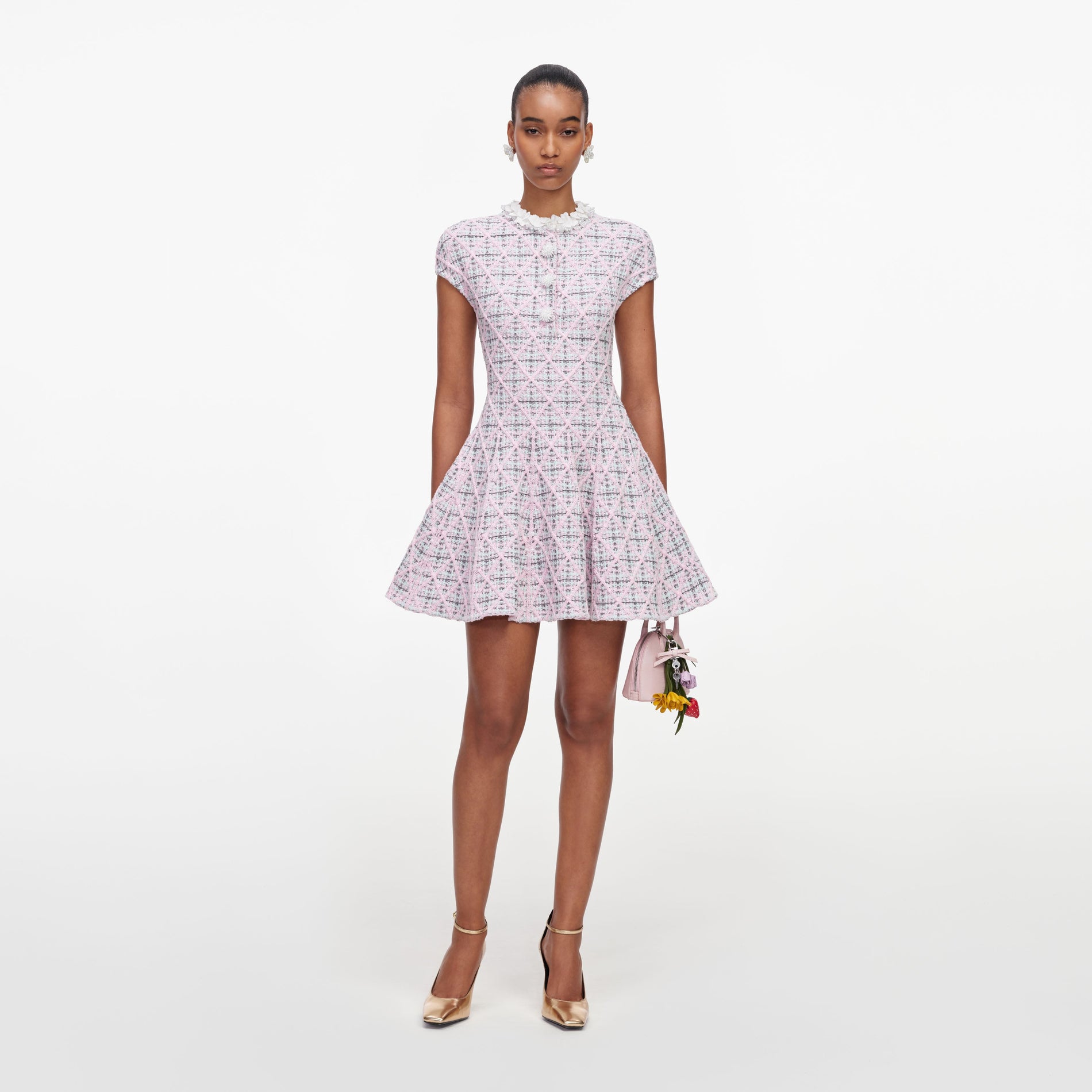 Model wearing a pink tweed dress with a ruffled collar, gold heels, and a pink handbag with floral charms