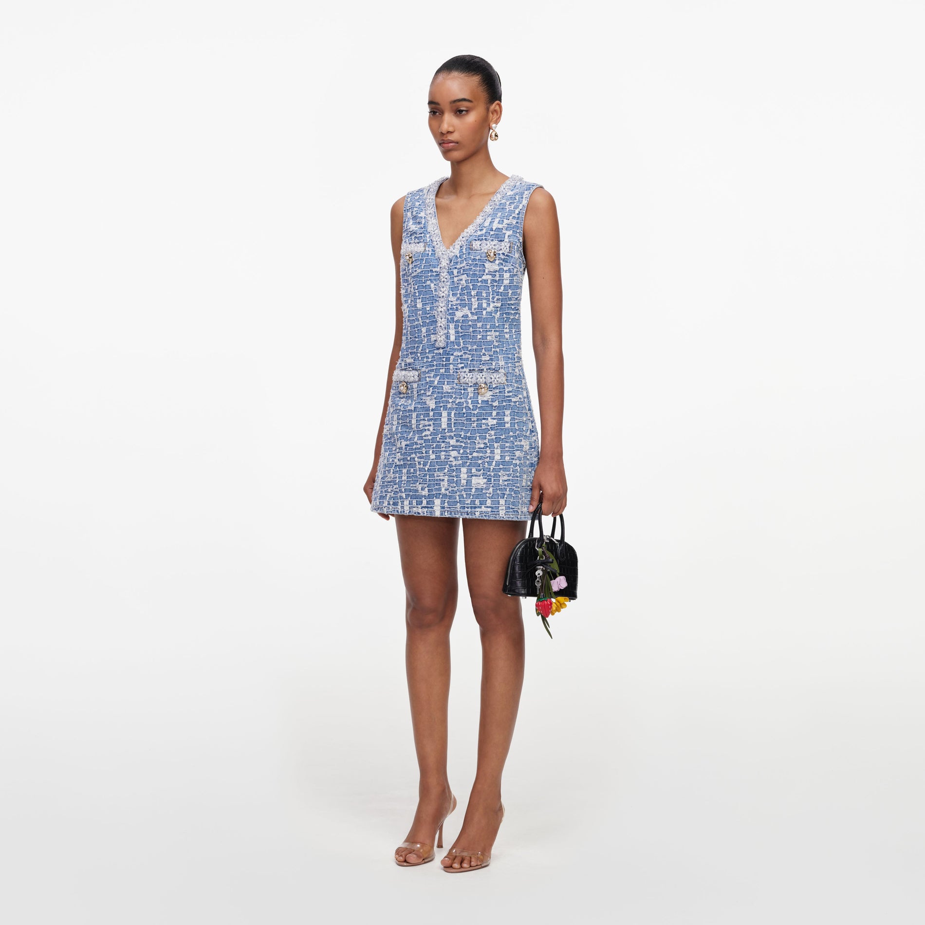 Woman in a blue and white tweed mini dress with gold buttons, holding a black handbag with floral charms