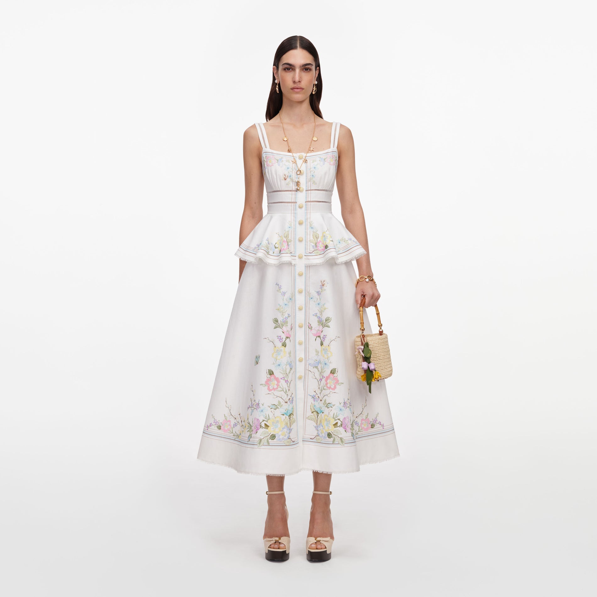 Model wearing a white floral embroidered dress with a peplum top and matching skirt, holding a straw bag