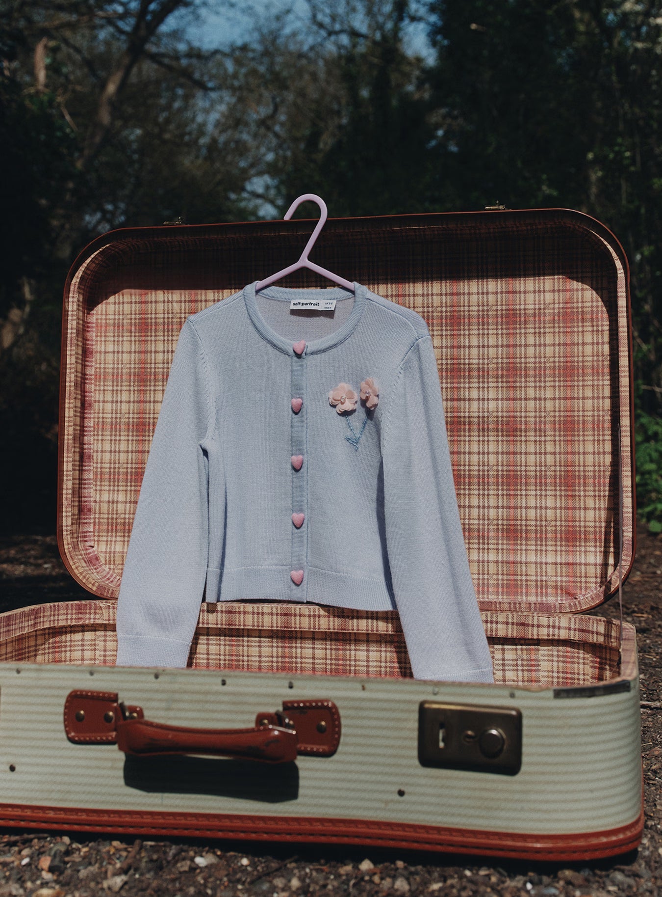 Light blue cardigan with pink heart buttons and flower details, hanging over an open vintage suitcase