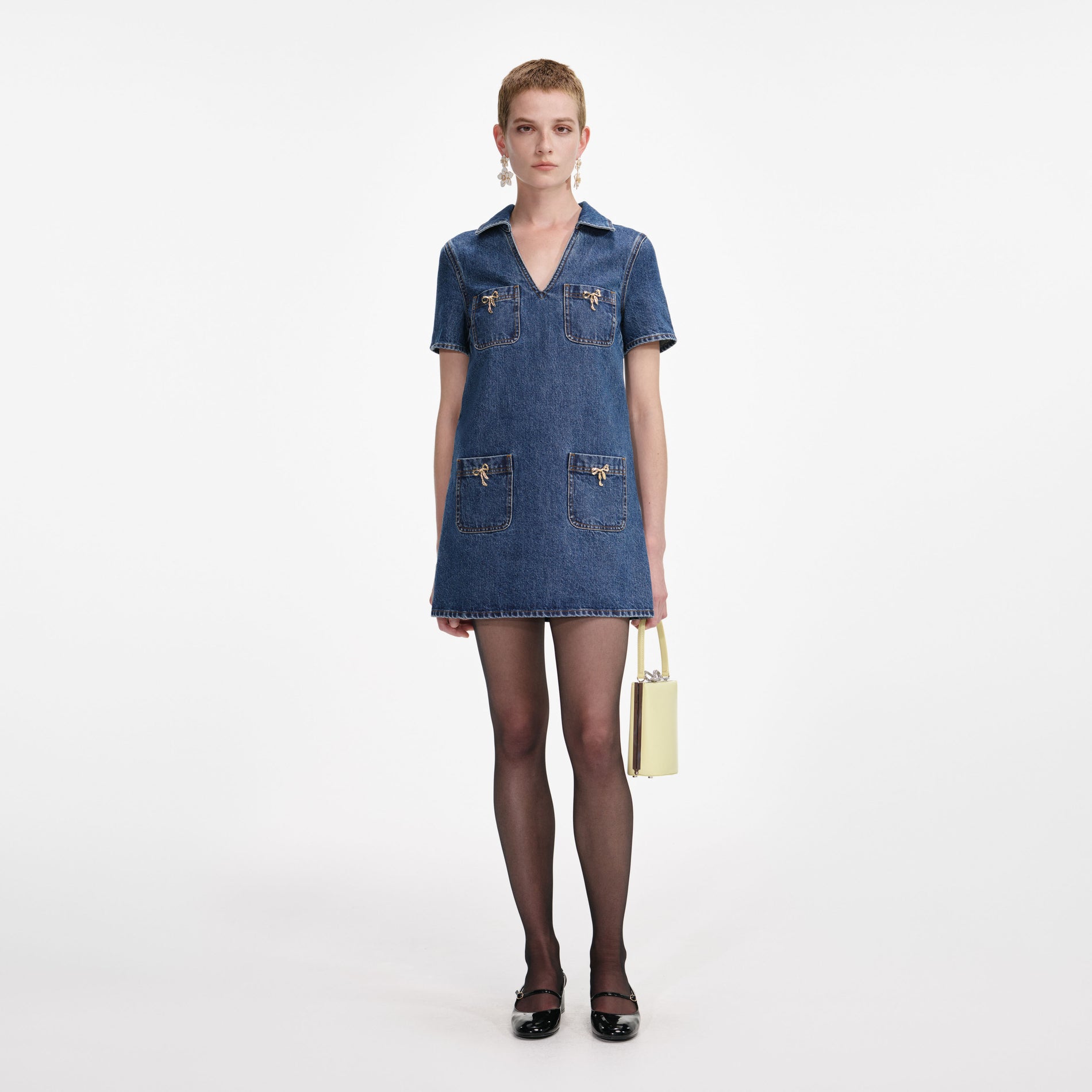 Woman wearing a denim mini dress with four pockets and gold bow details, black tights, and black patent Mary Jane shoes