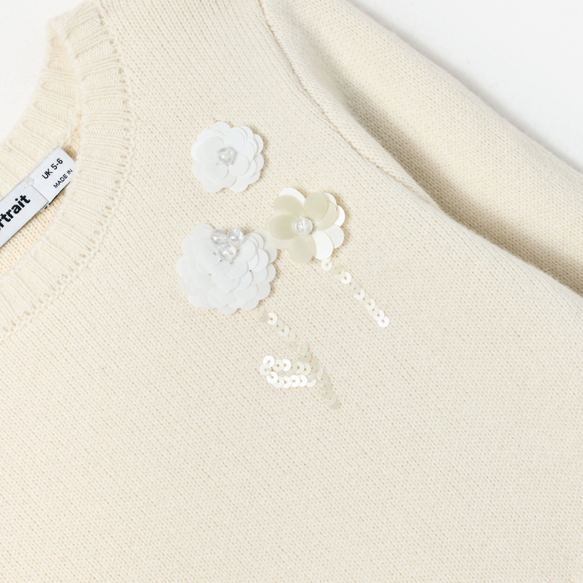 Close up of a cream knitted sweater with three white sequin flowers and a tag that reads UK 5-6