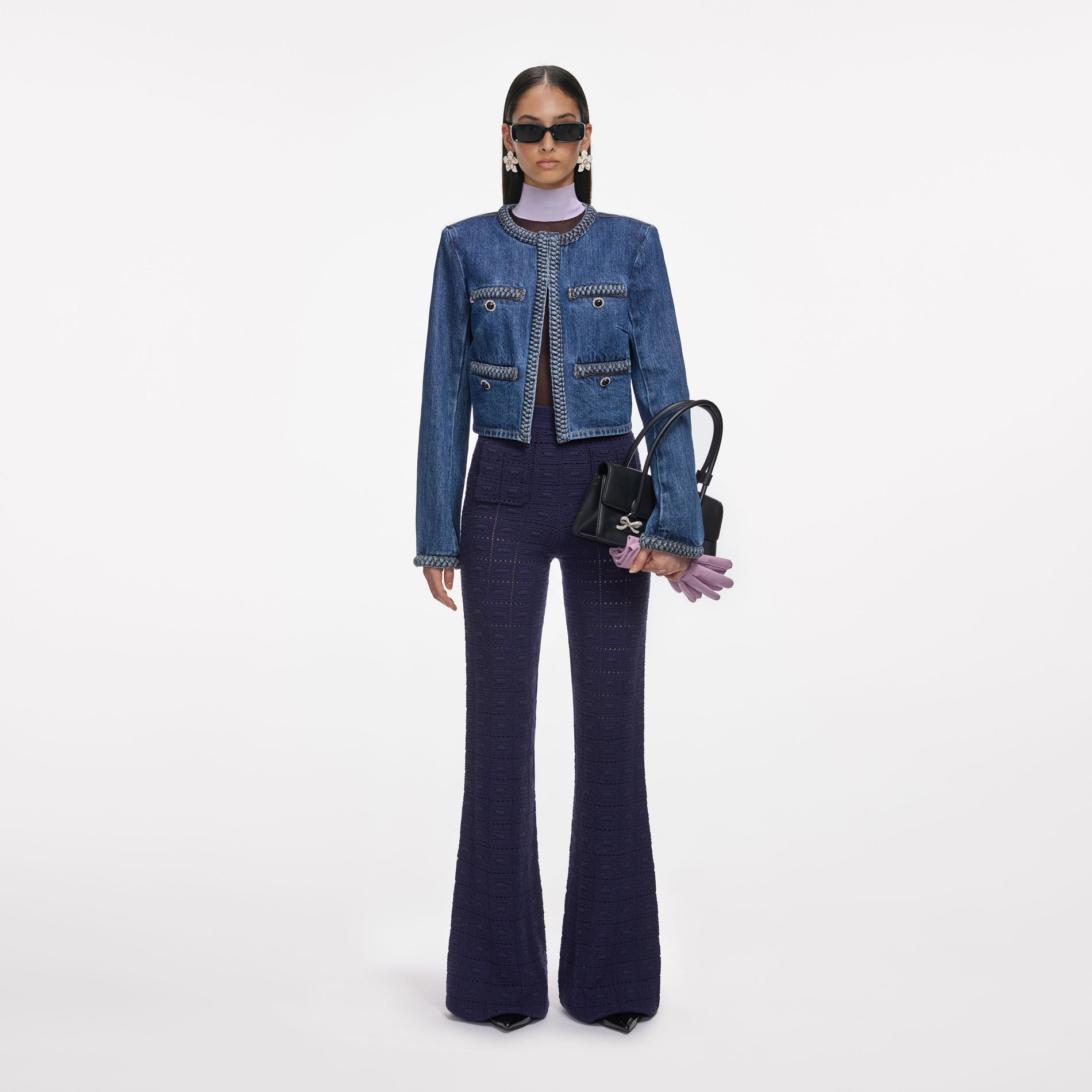 Woman in denim jacket, purple turtleneck, dark blue knit pants, sunglasses, holding black handbag and purple gloves
