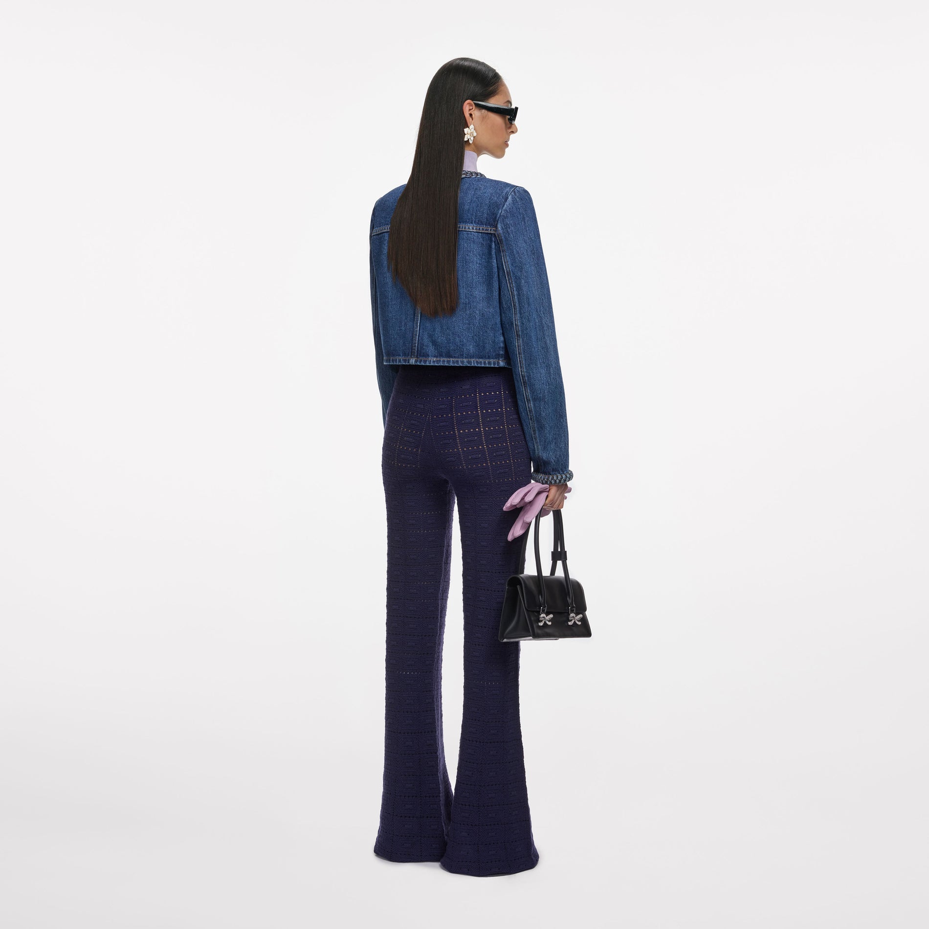 Woman from behind wearing a denim jacket, purple knit pants, black sunglasses, and holding a black handbag